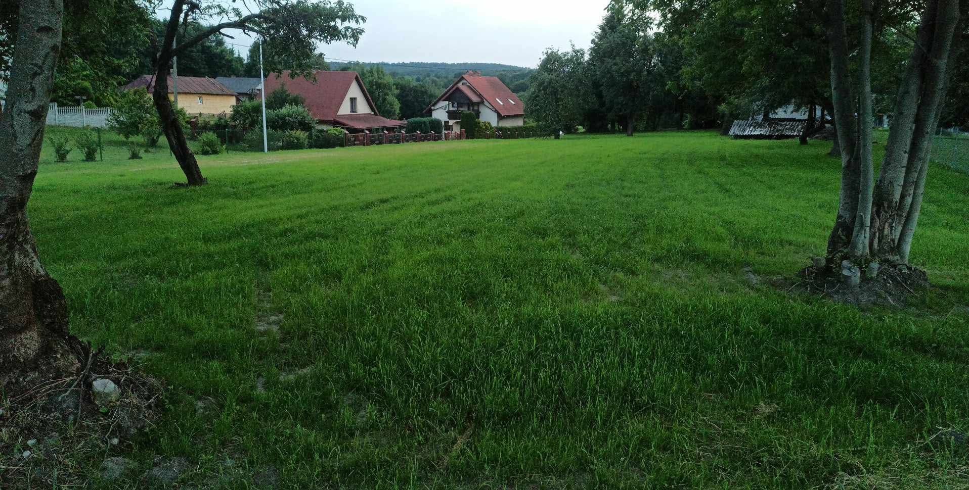 Rozległy trawnik przed domami jednorodzinnymi, z widocznymi drzewami po bokach kadru. Trawa jest świeżo skoszona, a w tle widać zabudowania mieszkalne.