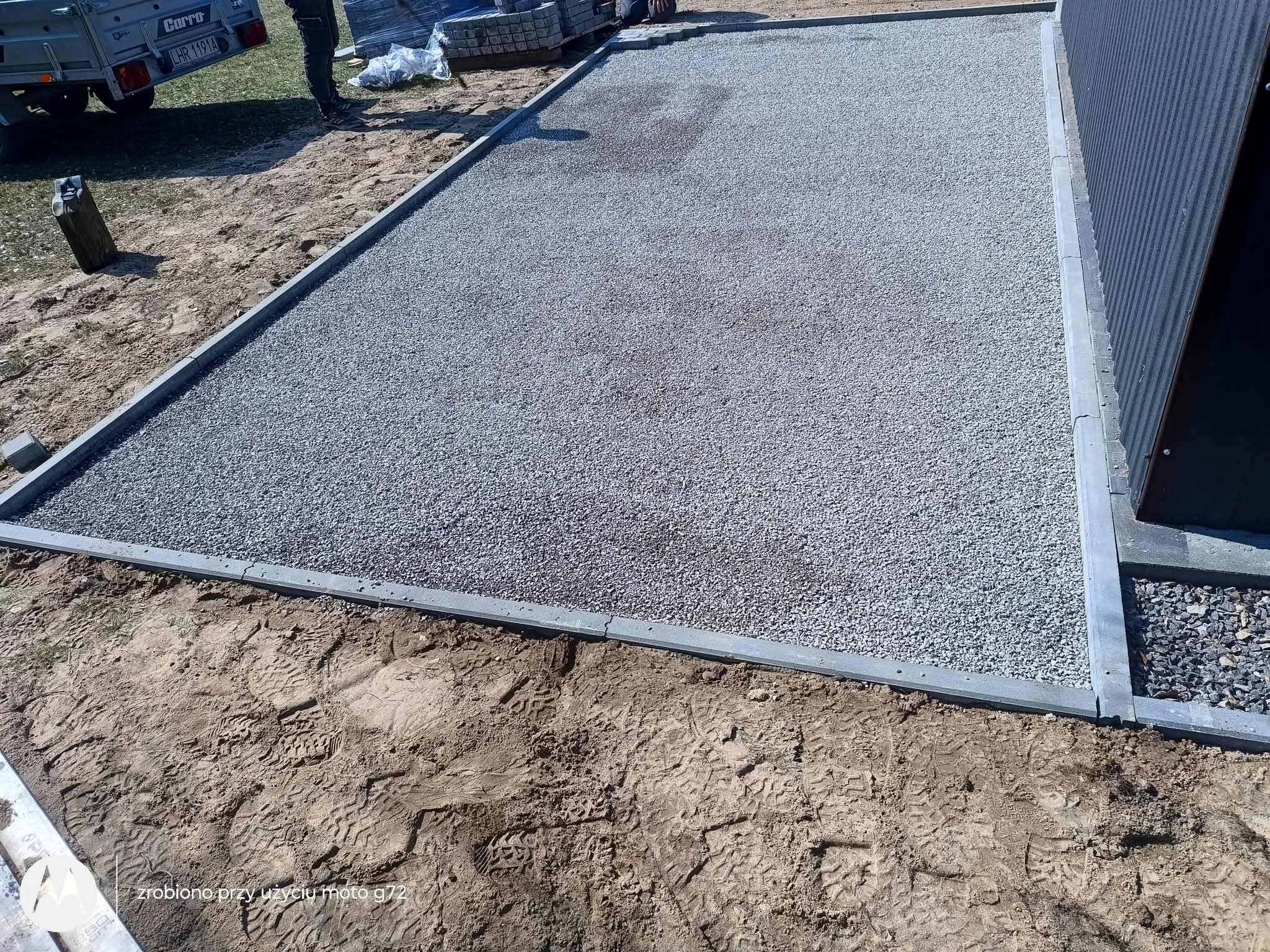 Utwardzony teren z szarą nawierzchnią i obrzeżami z kostki, widoczne ślady opon i butów na ziemi, przyczepa i paleta kostki w tle.