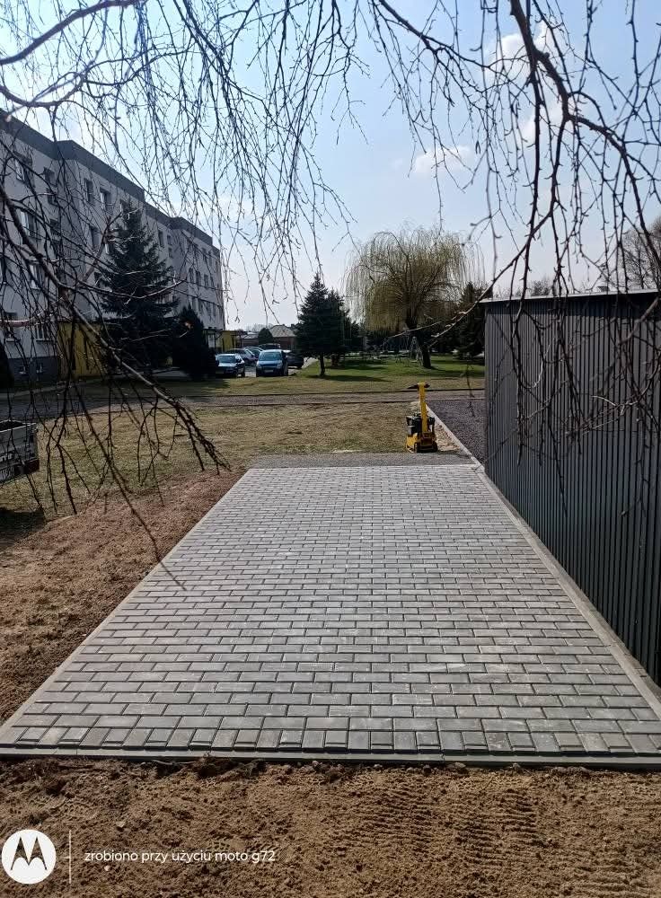 Ułożona kostka brukowa na terenie zielonym, widoczny budynek w tle i zagęszczarka wibracyjna. Kąt ujęcia z dołu podkreśla regularność wzoru kostki.