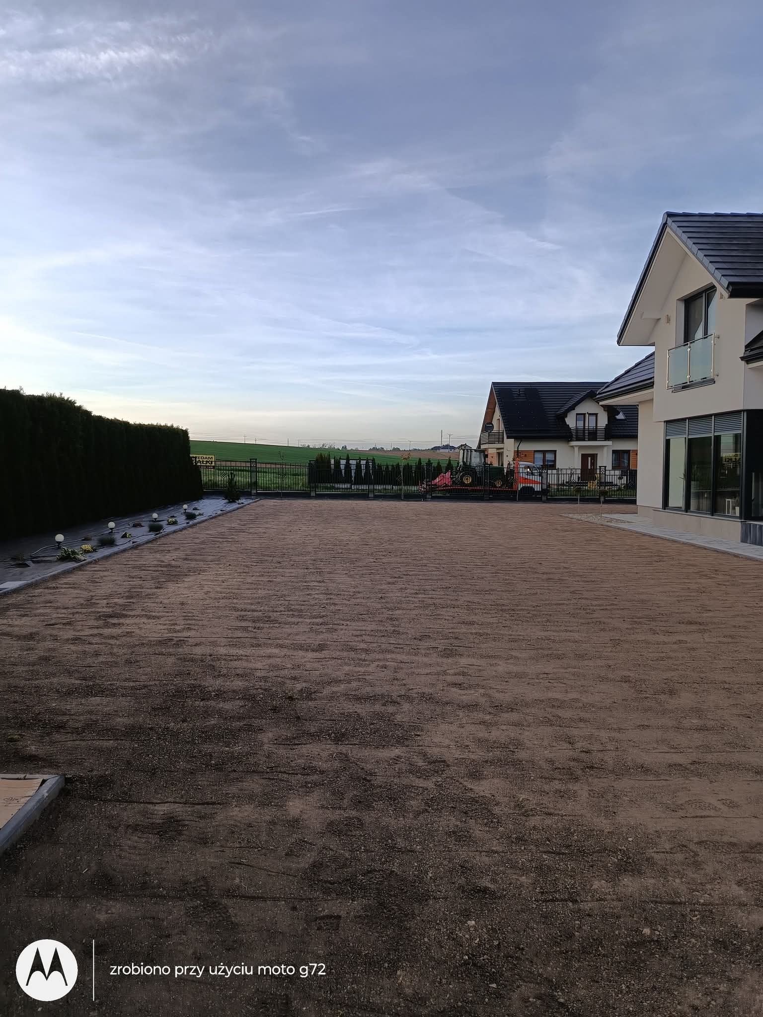 Przygotowany teren pod trawnik z rolki, widoczne ślady po wałowaniu. W tle domy jednorodzinne i traktor. Całość w pochmurny dzień.