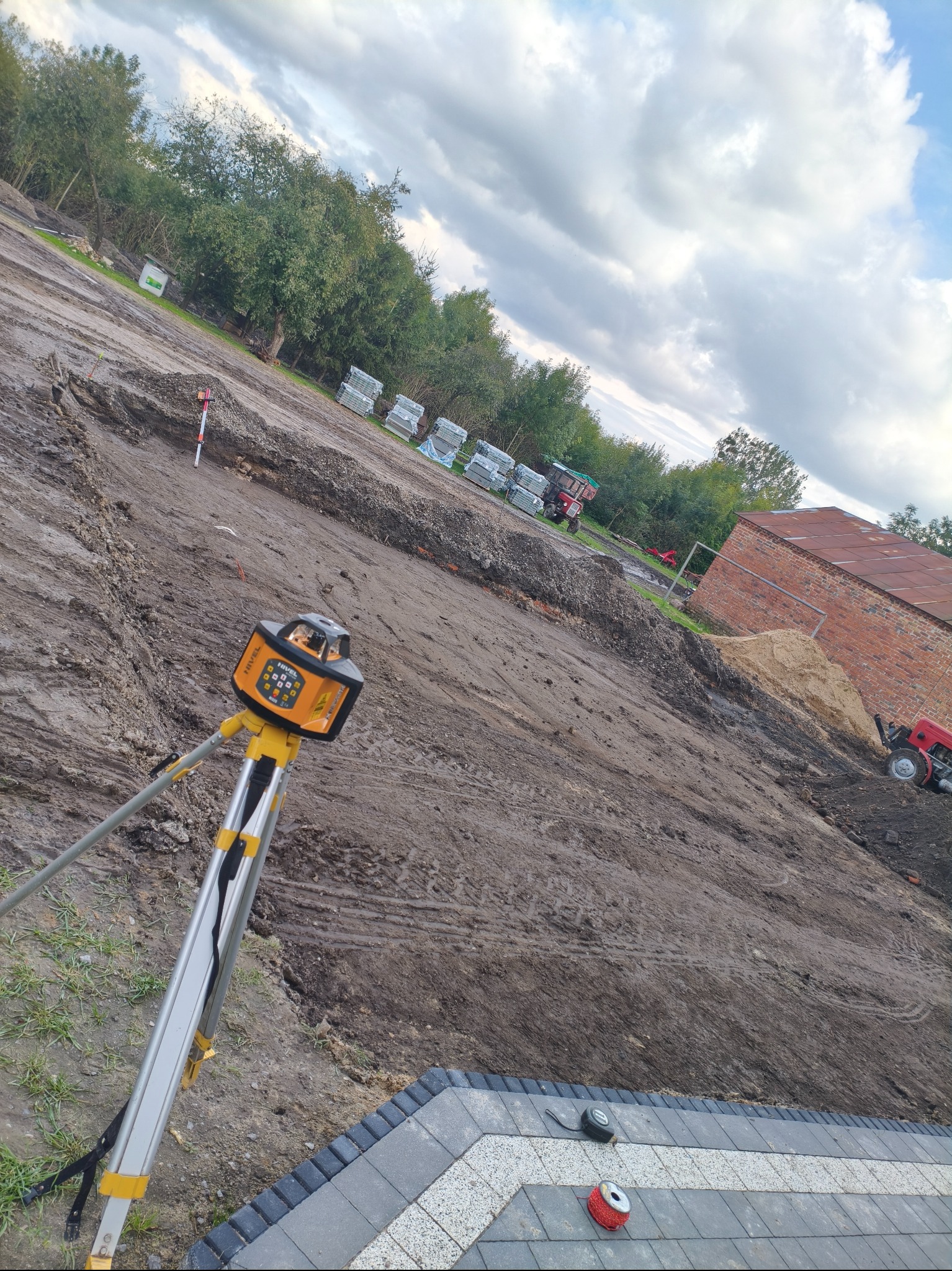 Niwelator laserowy na statywie, w tle przygotowany teren pod budowę z ułożoną kostką brukową. Widoczne narzędzia pomiarowe i maszyny budowlane.
