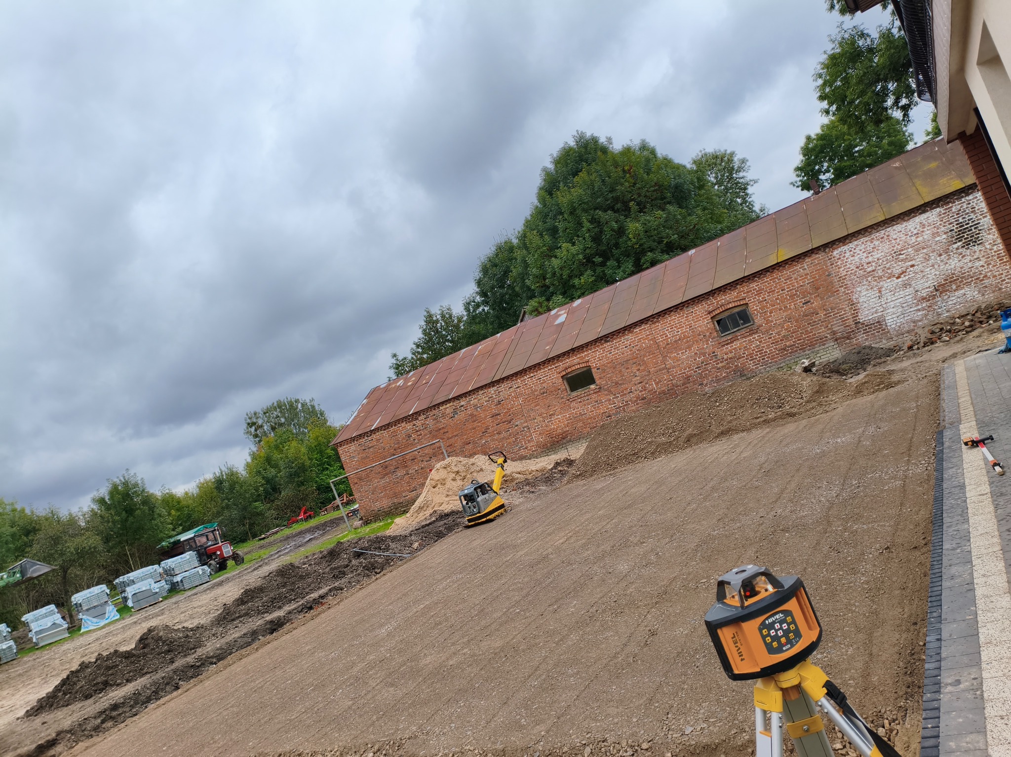 Przygotowanie terenu pod budowę: ubita ziemia, zagęszczarka, niwelator laserowy, w tle budynek gospodarczy z czerwonej cegły i palety z kostką brukową.