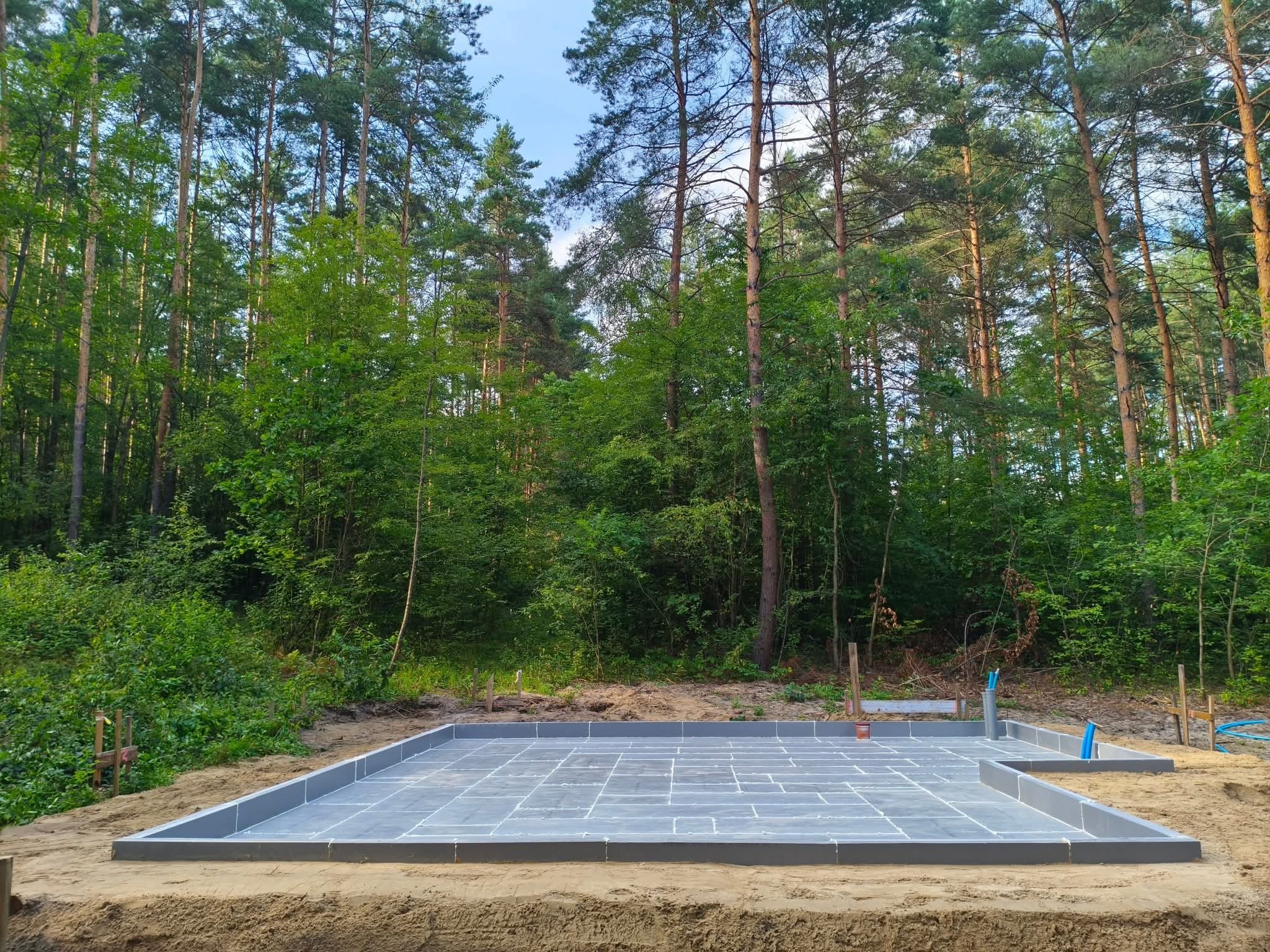 Gotowy fundament pod budowę domu w Adelina, otoczony lasem. Widoczne szare bloczki fundamentowe i piasek. Ujęcie z poziomu gruntu, podkreślające solidność konstrukcji.