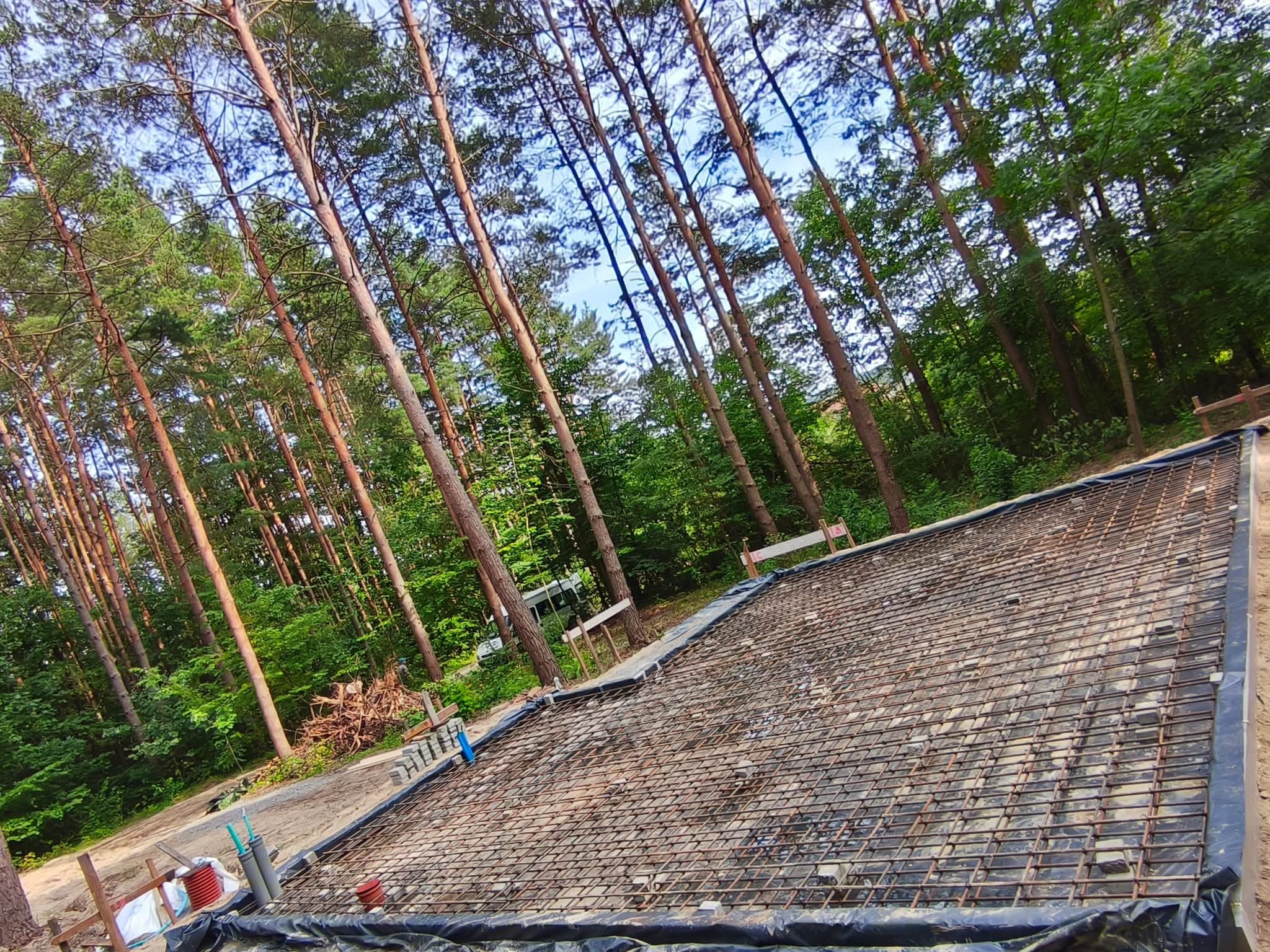 Zbrojenie pod płytę fundamentową na leśnej działce, widok z góry. Siatka zbrojeniowa ułożona na folii, otoczona szalunkiem. W tle wysokie drzewa iglaste i liściaste.