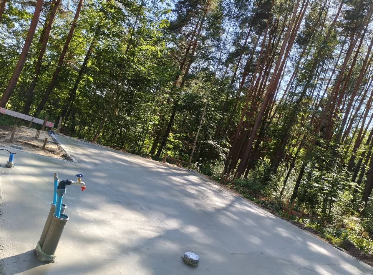 Wylana, gładka betonowa powierzchnia w otoczeniu drzew, z widocznymi elementami instalacji wodnej i drewnianymi szalunkami. Świeży beton w leśnej scenerii.