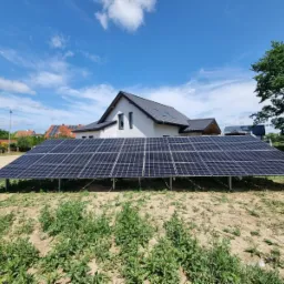 Instalacja paneli słonecznych na stelażu przed nowoczesnym domem z szarą dachówką w słoneczny dzień.