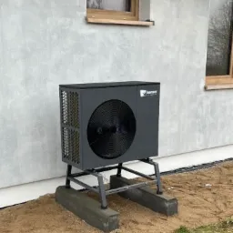 Szara pompa ciepła marki Kensol zainstalowana na metalowym stelażu, umieszczona na betonowych podstawach przed ścianą budynku z dwoma drewnianymi oknami.