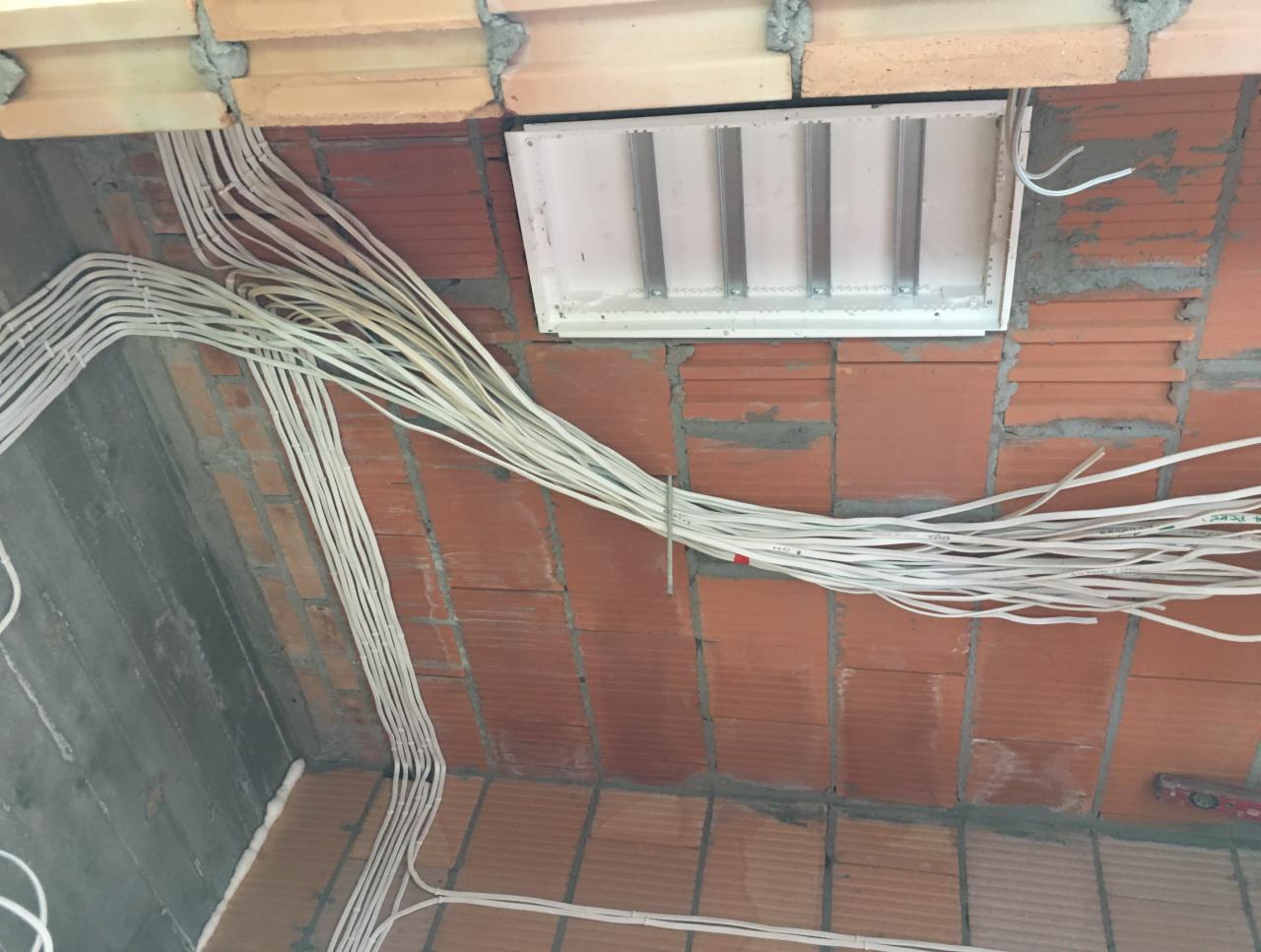 Rozprowadzenie przewodów elektrycznych w białej izolacji, mocowanych do sufitu z cegły, widoczna puszka rozdzielcza z metalowymi szynami.