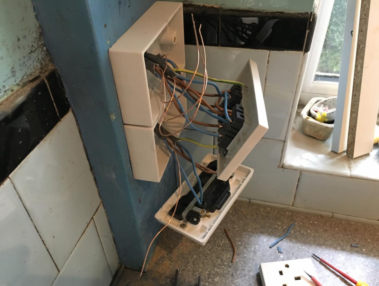 Otwarta puszka elektryczna z widocznymi przewodami w kolorach niebieskim, brązowym i zielono-żółtym, zamontowana na niebieskiej ścianie z białymi płytkami, obok widoczne narzędzia i fragment gniazdka.