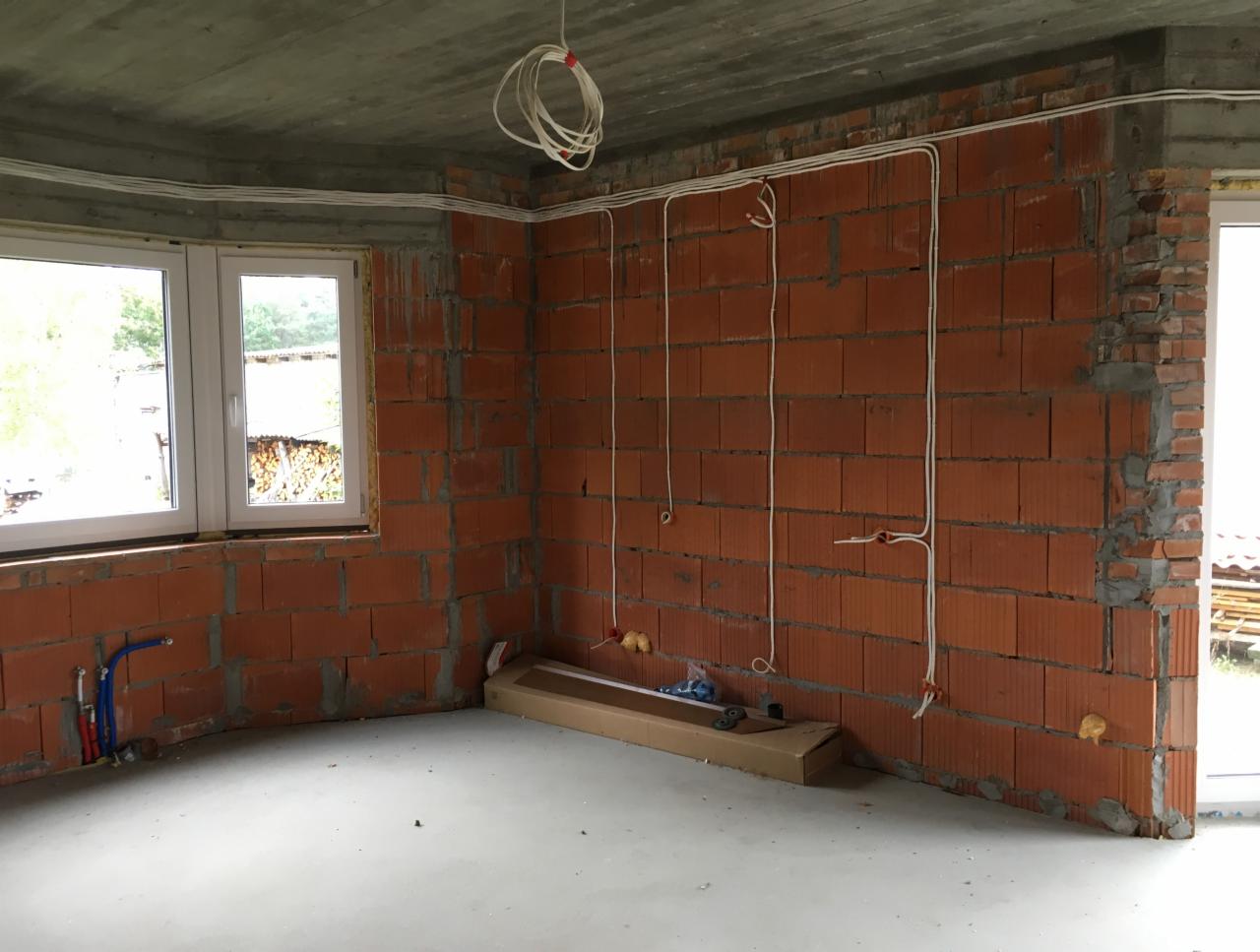 Wnętrze w stanie surowym w Warszawie z widoczną instalacją elektryczną poprowadzoną po ceglanych ścianach i pod sufitem, kable zwisają z sufitu, a pod oknem rury instalacji wodnej.
