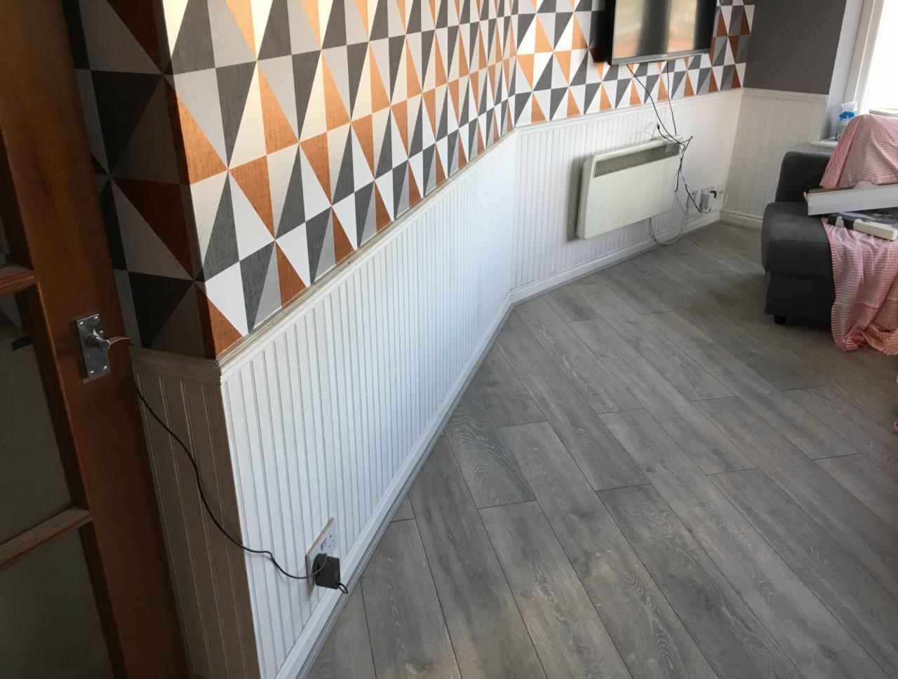 Salon po remoncie: ściana z geometryczną tapetą w odcieniach szarości, miedzi i bieli, dolna część ściany wykończona białą boazerią panelową, szara podłoga, grzejnik elektryczny na ścianie...