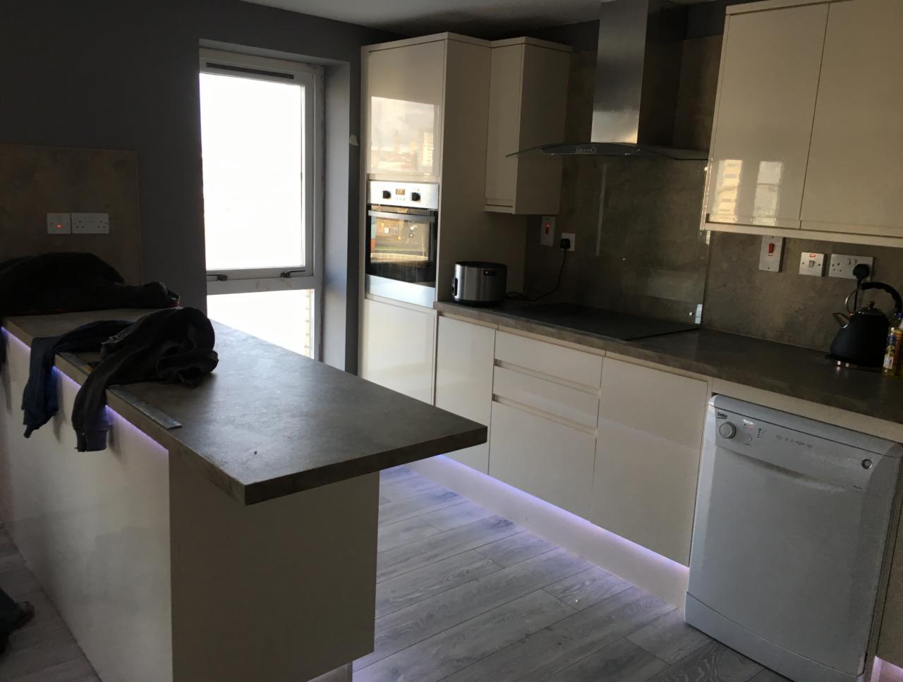 Nowoczesna kuchnia z białymi, błyszczącymi szafkami, szarym blatem i podświetleniem LED pod szafkami i wyspą kuchenną. Widoczne gniazdka elektryczne na ścianie i płyta indukcyjna na blacie.