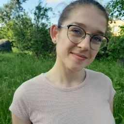 Uśmiechnięta młoda kobieta w okularach, ubrana w prążkowaną koszulkę, pozuje na tle zielonej trawy i drzew w słoneczny dzień.