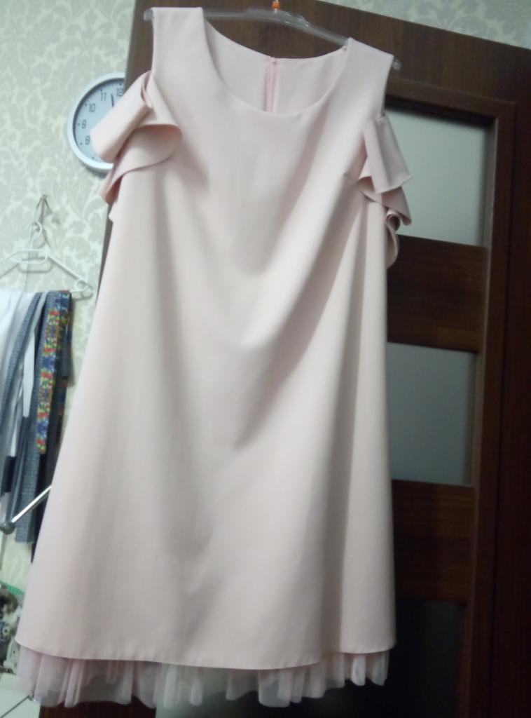 Jasnoróżowa sukienka na wieszaku z falbankami na ramionach i tiulowym podszyciem, wisząca na tle wzorzystej tapety i drewnianych drzwi.