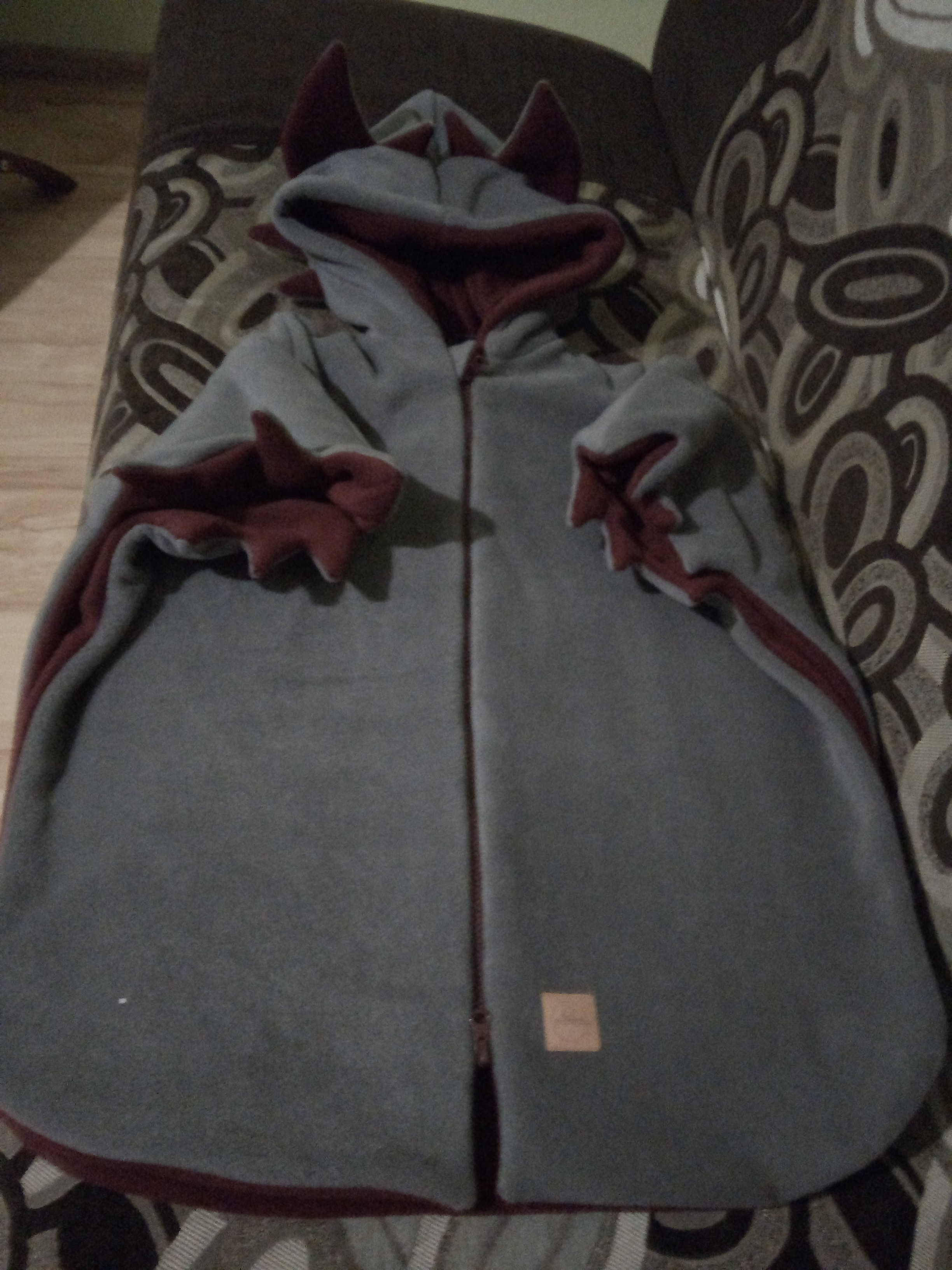 Rozłożona, szara bluza dziecięca z kapturem i zdobieniami w kształcie rogów, wykończona bordową lamówką, leżąca na wzorzystej kanapie.