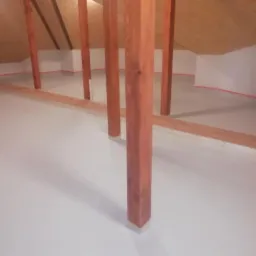 Jasnoszara, gładka posadzka w surowym wnętrzu poddasza z drewnianą konstrukcją, zabezpieczona taśmą malarską przy ścianach. Widoczne drewniane słupy.