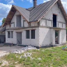 Dwukondygnacyjny dom murowany w trakcie budowy, z widoczną konstrukcją dachu pokrytą papą i kominem, ściany z białych bloczków, na ziemi leżą resztki materiałów budowlanych, tło: błękitne niebo...