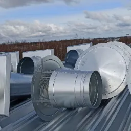 Elementy systemu wentylacyjnego z blachy ocynkowanej, w tym kanały okrągłe i kwadratowe, oraz okrągłe zakończenia z siatką zabezpieczającą, ułożone na dachu z blachy trapezowej, na tle pochmurnego...