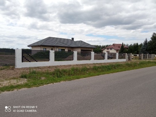 Długa, pozioma, nowoczesna linia ogrodzenia z betonowymi słupkami i czarnymi, poziomymi przęsłami, oddzielająca posesję od drogi asfaltowej w pochmurny dzień.