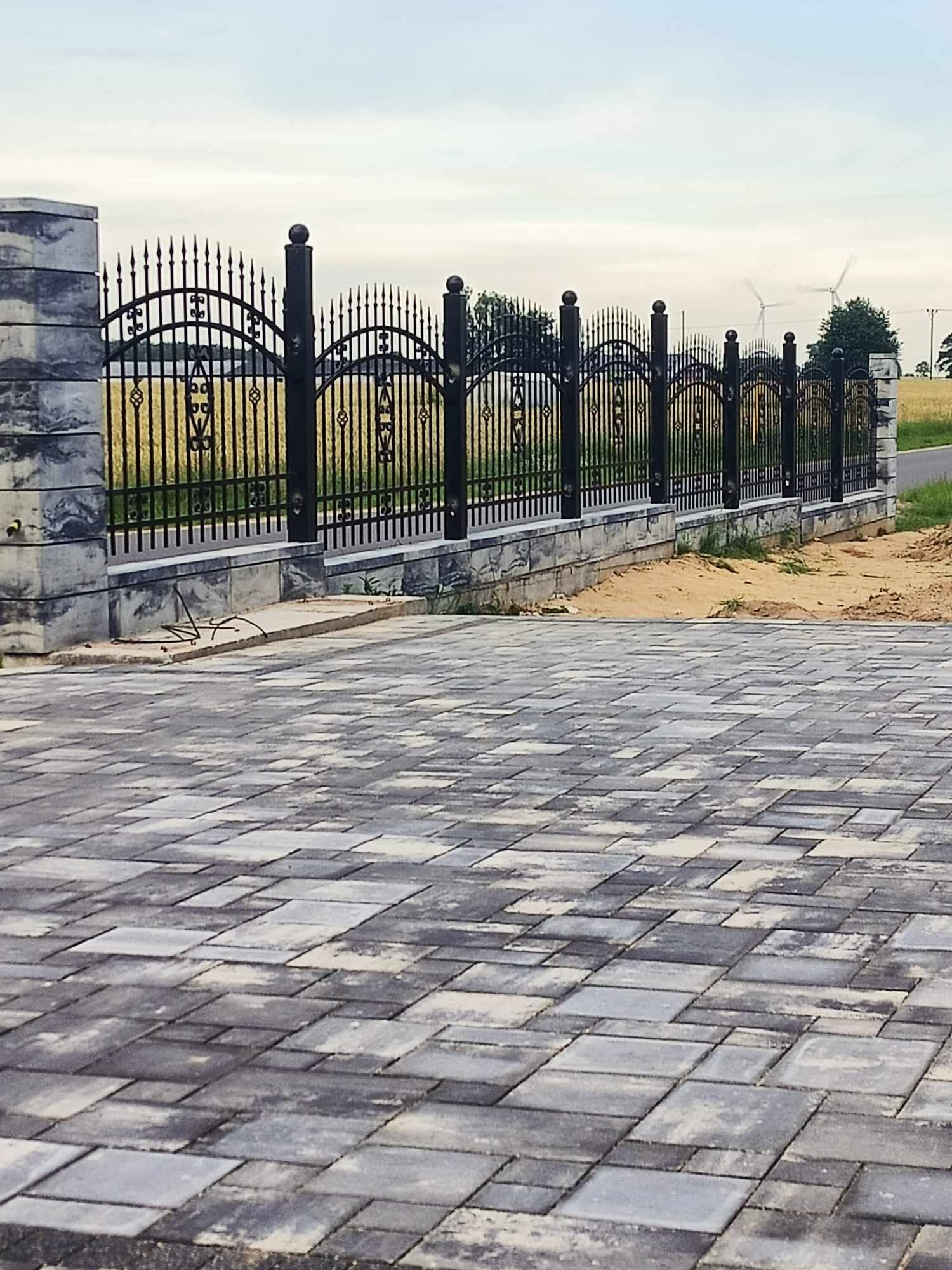 Czarne, kute ogrodzenie z ozdobnymi elementami, osadzone na niskim murku z szarych, ciosanych kamieni; w tle pole i turbina wiatrowa, na pierwszym planie szara kostka brukowa.