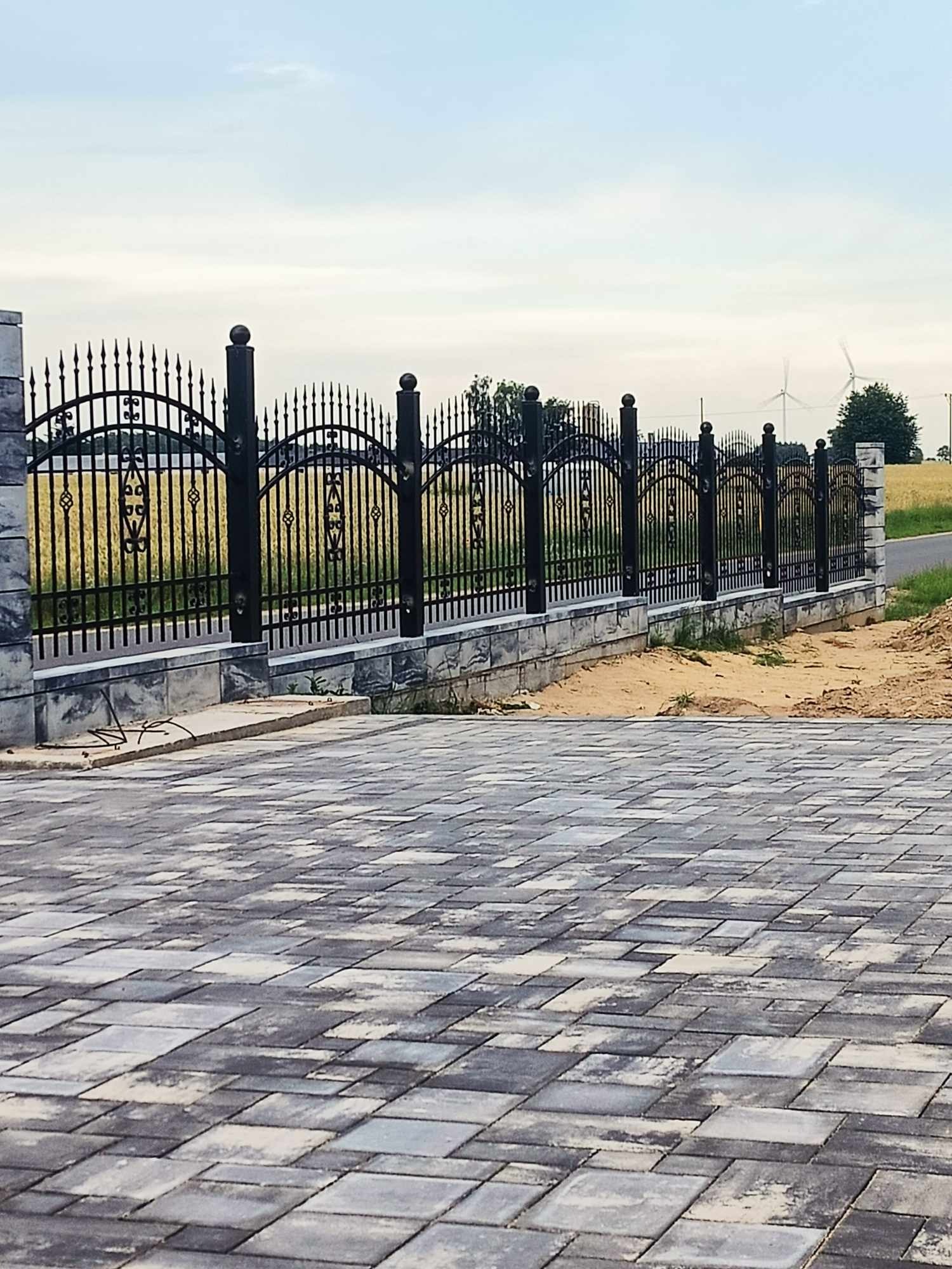 Czarny, metalowy płot z dekoracyjnymi elementami, osadzony na niskiej, kamiennej podmurówce, widok z brukowanego podjazdu, pole w tle z turbinami wiatrowymi.