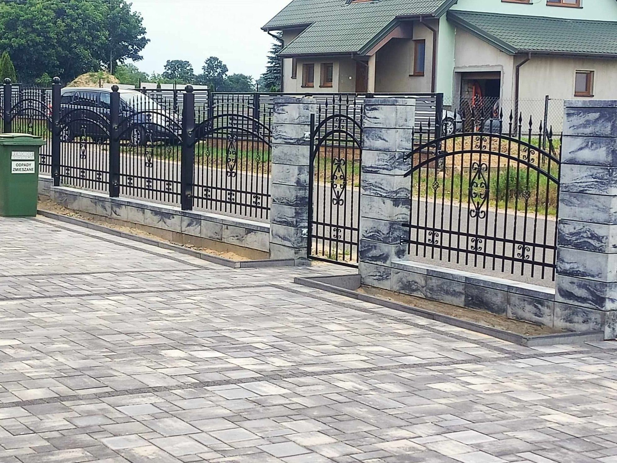 Nowoczesne ogrodzenie z czarnych, metalowych przęseł i szarych, betonowych słupków, oddzielające posesję z kostki brukowej od ulicy, z widocznym zielonym pojemnikiem na śmieci.