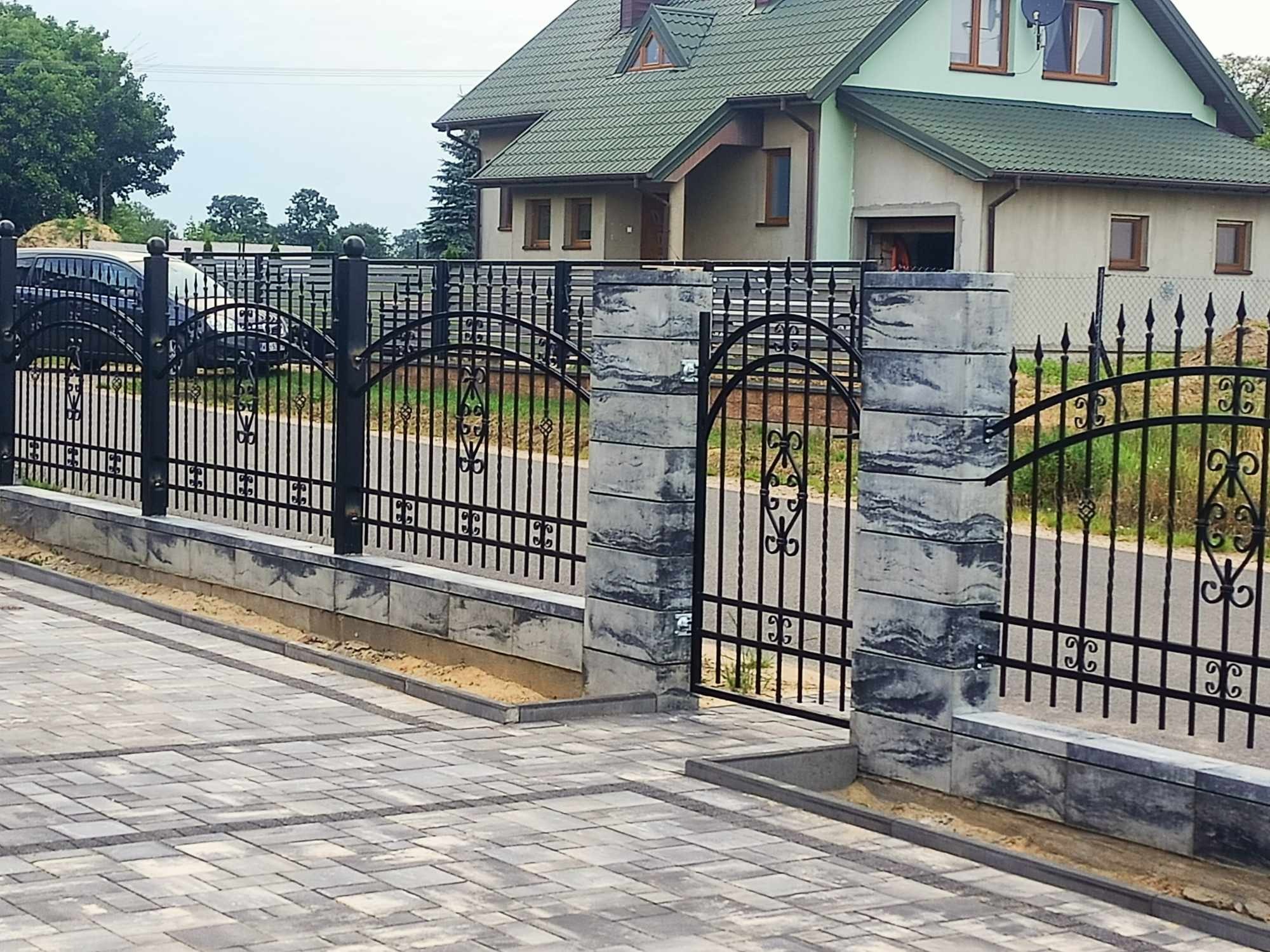 Czarna, kuta brama wjazdowa z dekoracyjnymi elementami, osadzona na betonowych słupkach z efektem marmuru, prowadząca do posesji z kostką brukową i domem w tle.