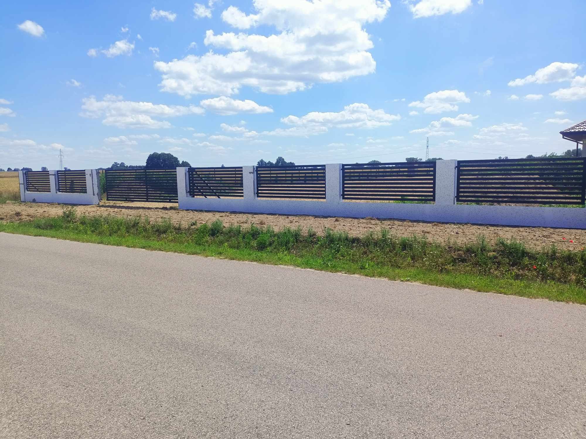 Nowoczesne ogrodzenie z poziomych, ciemnych paneli metalowych osadzonych w białych, murowanych słupkach i podmurówce, widok z perspektywy drogi na tle pola i błękitnego nieba z chmurami.