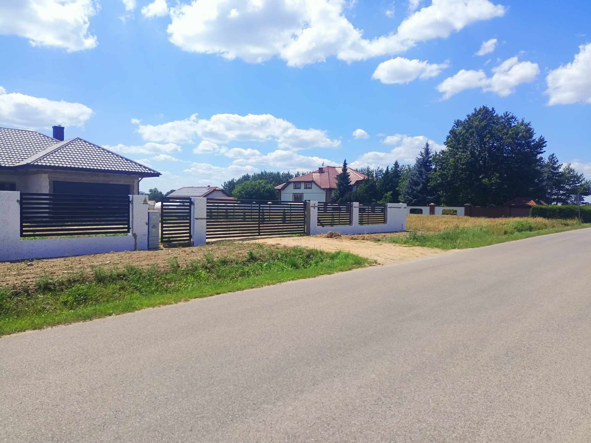 Nowoczesne ogrodzenie posesji z poziomych, czarnych profili na białych, murowanych słupkach, widok z poziomu ulicy w słoneczny dzień.