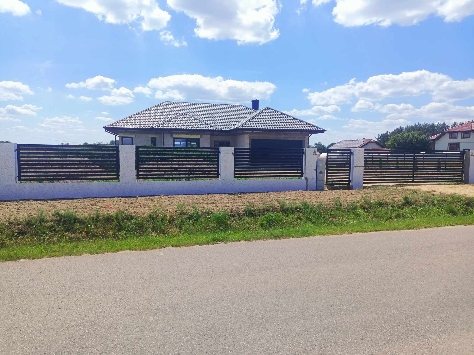 Nowoczesne ogrodzenie posesji z poziomych, czarnych elementów w Białkowie, z bramą wjazdową i furtką, na tle domu jednorodzinnego i błękitnego nieba.