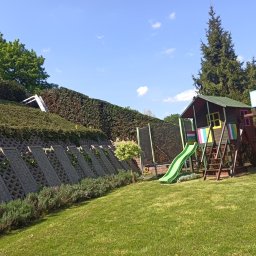 ZZ-UP Zdzisław Zebranowski - Utrzymany trawnik z domkiem dziecięcym i trampoliną, na zboczu wzmocnionym betonowymi elementami i obsadzonym roślinnością. Niebo z delikatnymi chmurami w tle.