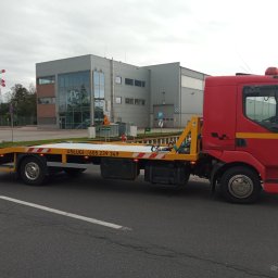 ZZ-UP Zdzisław Zebranowski - Czerwona laweta z żółtą platformą na szosie, z numerem telefonu i budynkiem w tle. Widoczny znak stop i przejazd kolejowy. Pochmurne niebo.