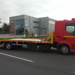 Czerwona laweta z żółtą platformą na szosie, z numerem telefonu i budynkiem w tle. Widoczny znak stop i przejazd kolejowy. Pochmurne niebo.