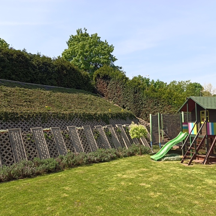 Utrzymany trawnik z elementami architektury ogrodowej: ażurowe panele, skarpa z roślinnością, plac zabaw w słoneczny dzień.