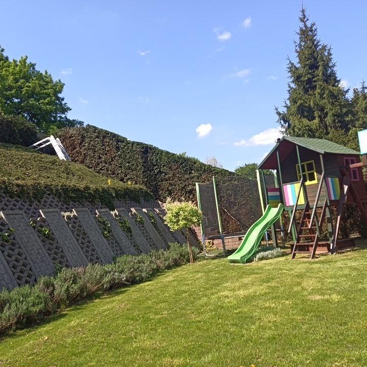 Utrzymany trawnik z domkiem dziecięcym i trampoliną, na zboczu wzmocnionym betonowymi elementami i obsadzonym roślinnością. Niebo z delikatnymi chmurami w tle.