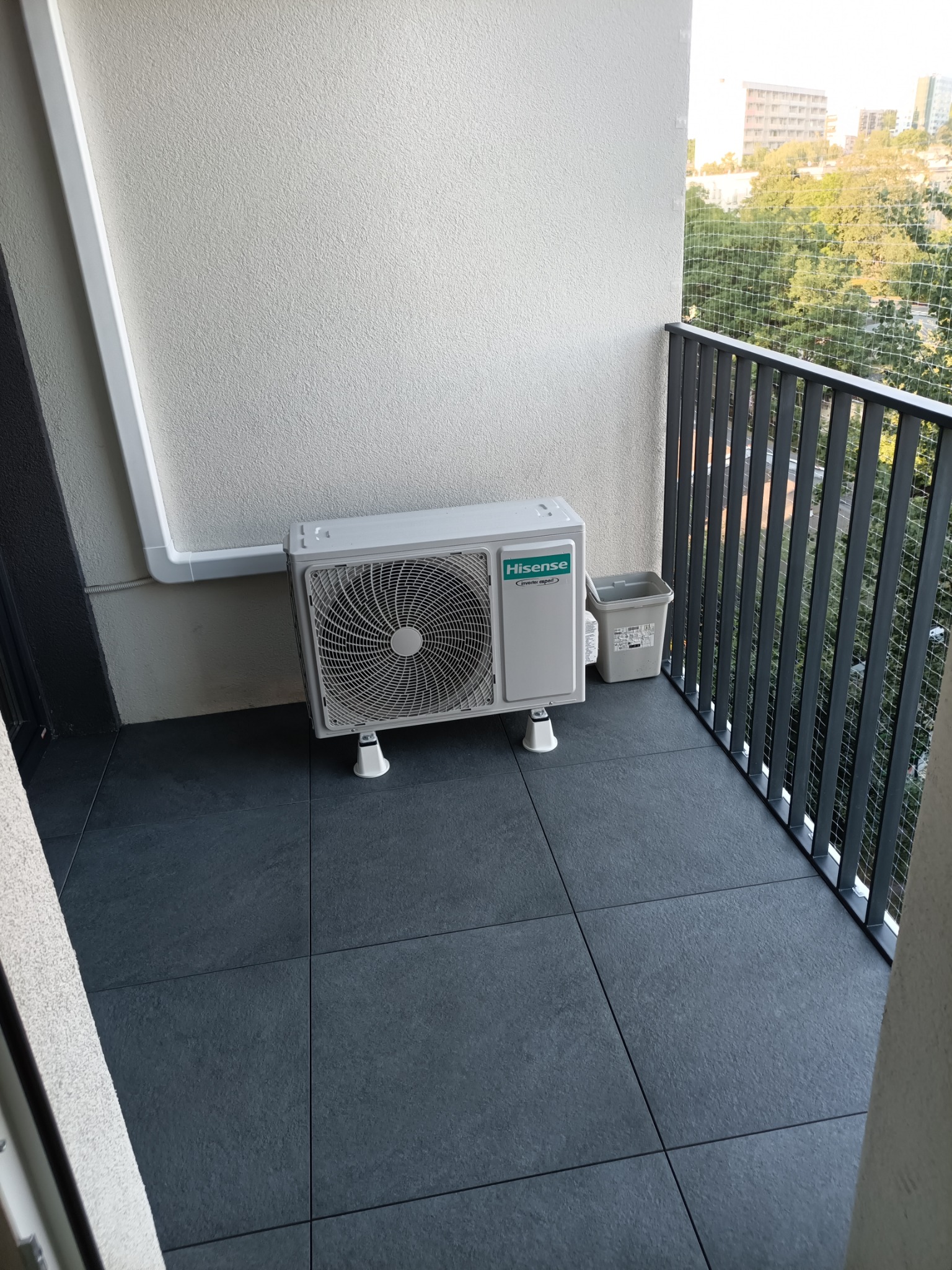Kompaktowy klimatyzator Hisense na balkonie z szarymi płytkami i metalową balustradą. Widok na zieleń miejską przez siatkę ochronną.