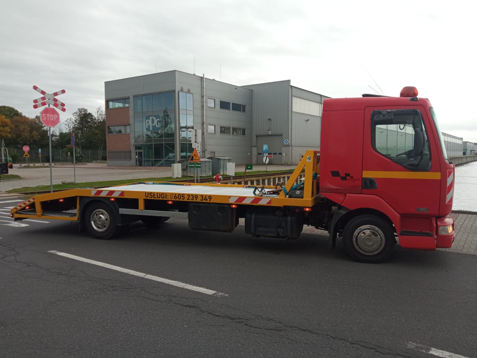 Czerwona laweta z żółtą platformą na szosie, z numerem telefonu i budynkiem w tle. Widoczny znak stop i przejazd kolejowy. Pochmurne niebo.