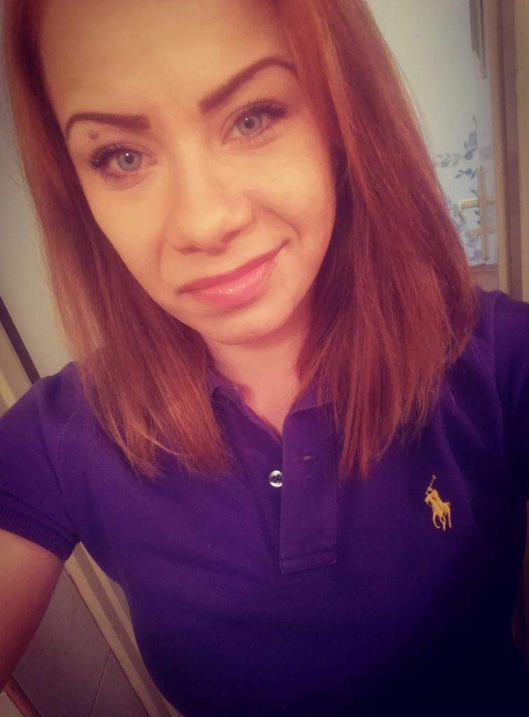 Portret kobiety o rudych włosach w fioletowej koszulce polo z logo, uśmiechnięta, z widocznym fragmentem łazienki w tle.