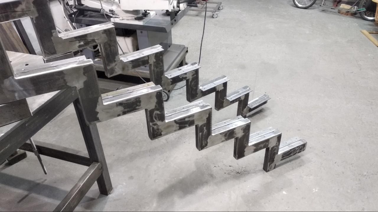 Surowe, metalowe schody z profili stalowych w trakcie produkcji, widoczne ślady po spawaniu, szlifowaniu i obróbce. W tle maszyny warsztatowe.