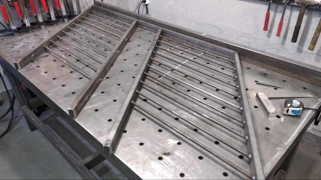 Metalowa konstrukcja z prętów na perforowanym stole spawalniczym, widoczne narzędzia pomiarowe i młotki na ścianie.