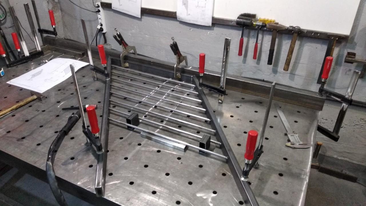 Metalowa konstrukcja kratowa w trakcie montażu na perforowanym stole warsztatowym, zabezpieczona ściskami stolarskimi z czerwonymi uchwytami, obok rysunek techniczny i narzędzia.