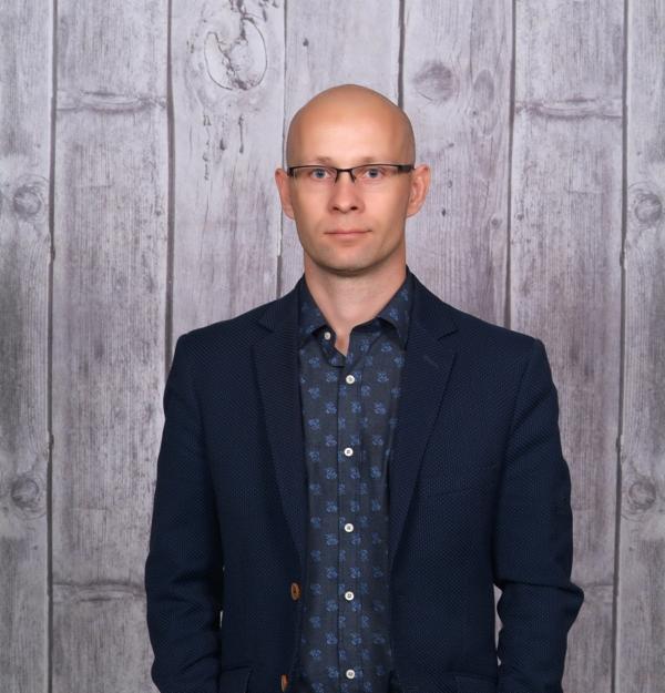 Portret mężczyzny w okularach, granatowej marynarce i koszuli w drobny wzór na tle drewnianej ściany.