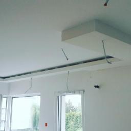 Nowaprzestrzen - Wnętrze w trakcie prac wykończeniowych: sufit z elementami z płyty karton gips, widoczne kable elektryczne, dwa okna z widokiem na zieleń.