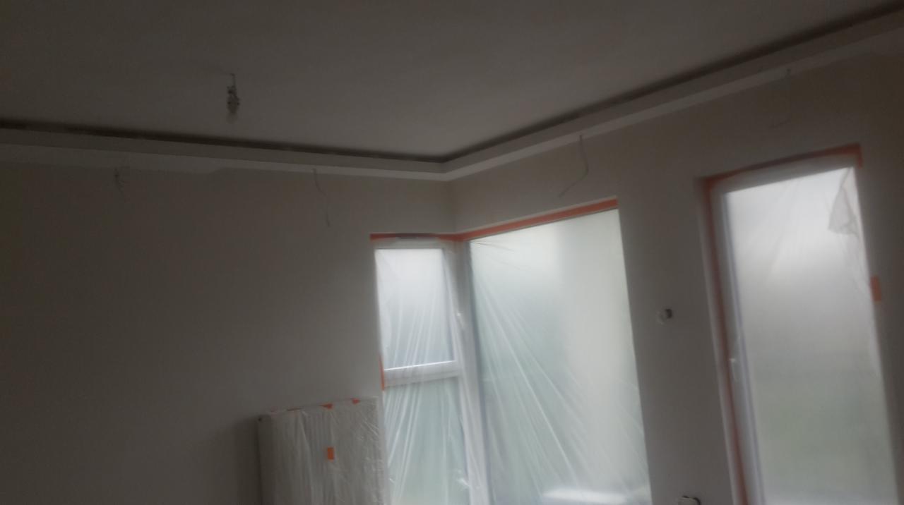 Przygotowanie do malowania w Warszawie: Pokój z zabezpieczonymi folią oknami i grzejnikiem, świeżo pomalowane ściany na szaro, widoczne listwy przysufitowe.