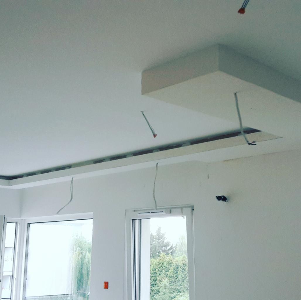 Wnętrze w trakcie prac wykończeniowych: sufit z elementami z płyty karton gips, widoczne kable elektryczne, dwa okna z widokiem na zieleń.