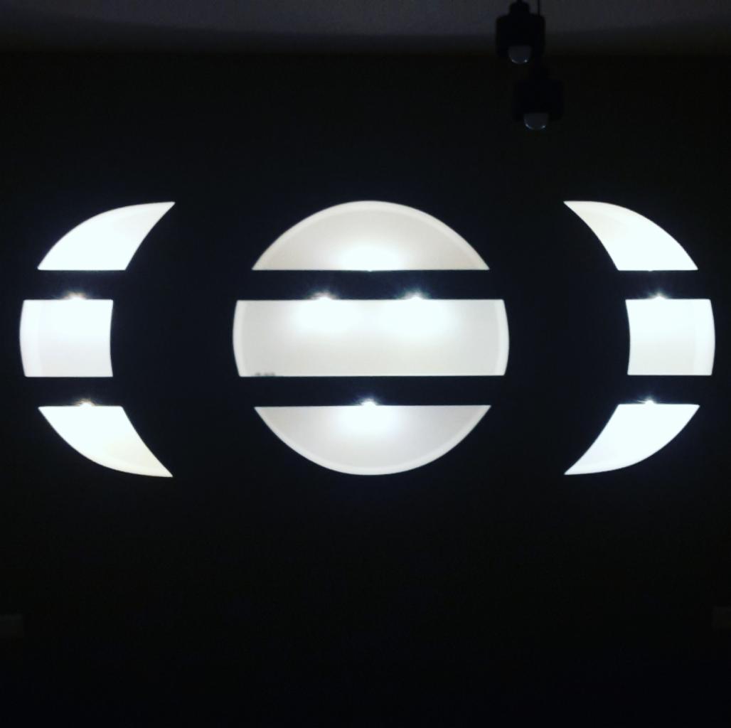 Podświetlana dekoracja ścienna w kształcie trzech okręgów, przeciętych poziomymi pasami, na ciemnym tle ściany.