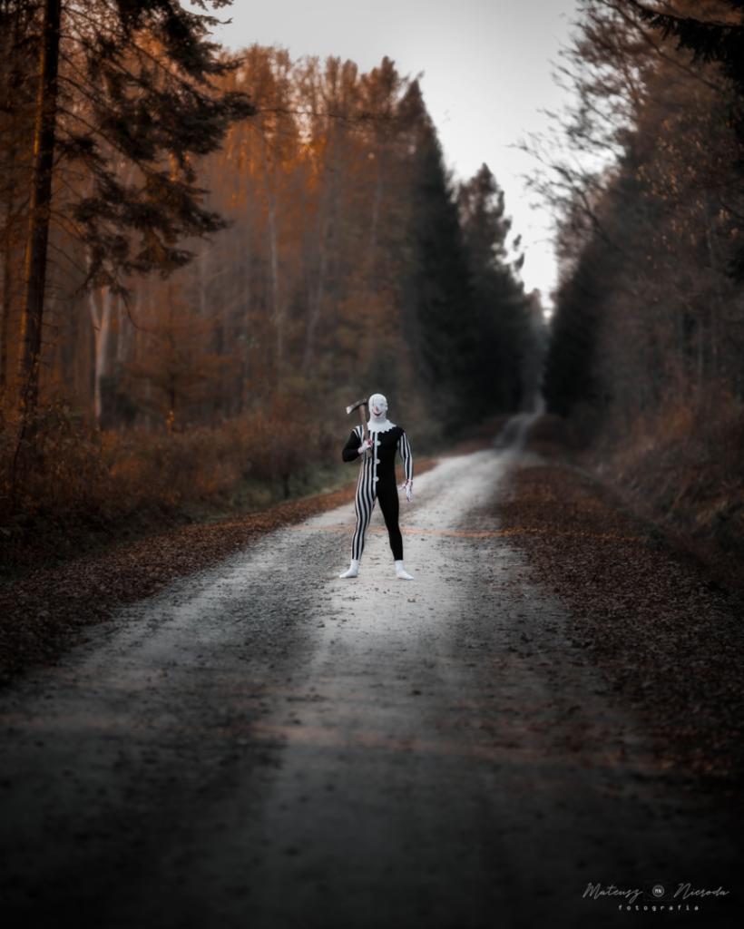 Osoba w kostiumie klauna w paski, z toporem na ramieniu, stoi na leśnej drodze, otoczona jesiennymi drzewami.