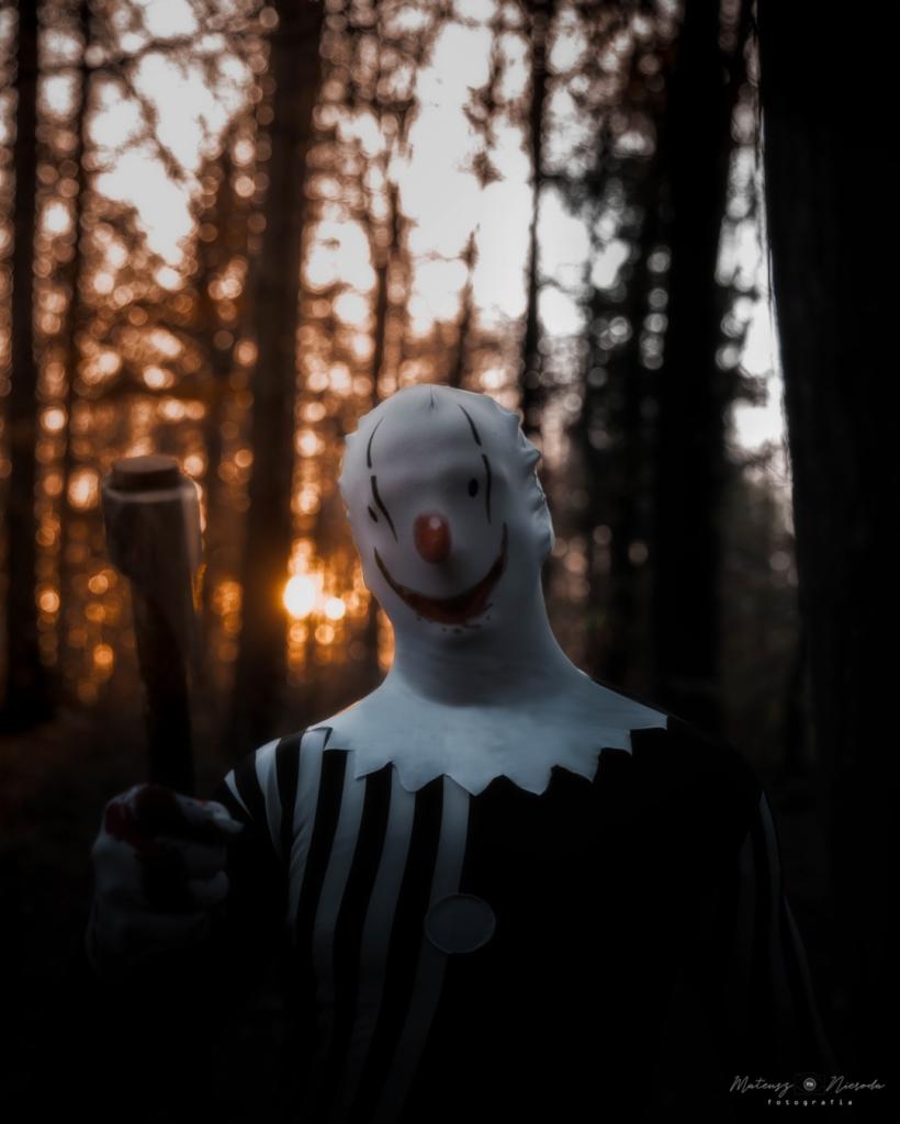 Mroczna fotografia klauna w masce z toporem w lesie, oświetlonego zachodzącym słońcem.