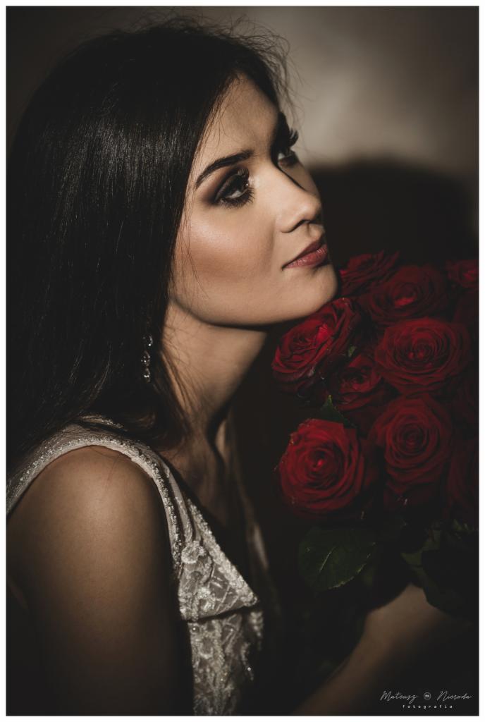 Portret kobiety z ciemnymi włosami i bukietem czerwonych róż, spoglądającej w górę, w sukni z koronkowym wykończeniem.