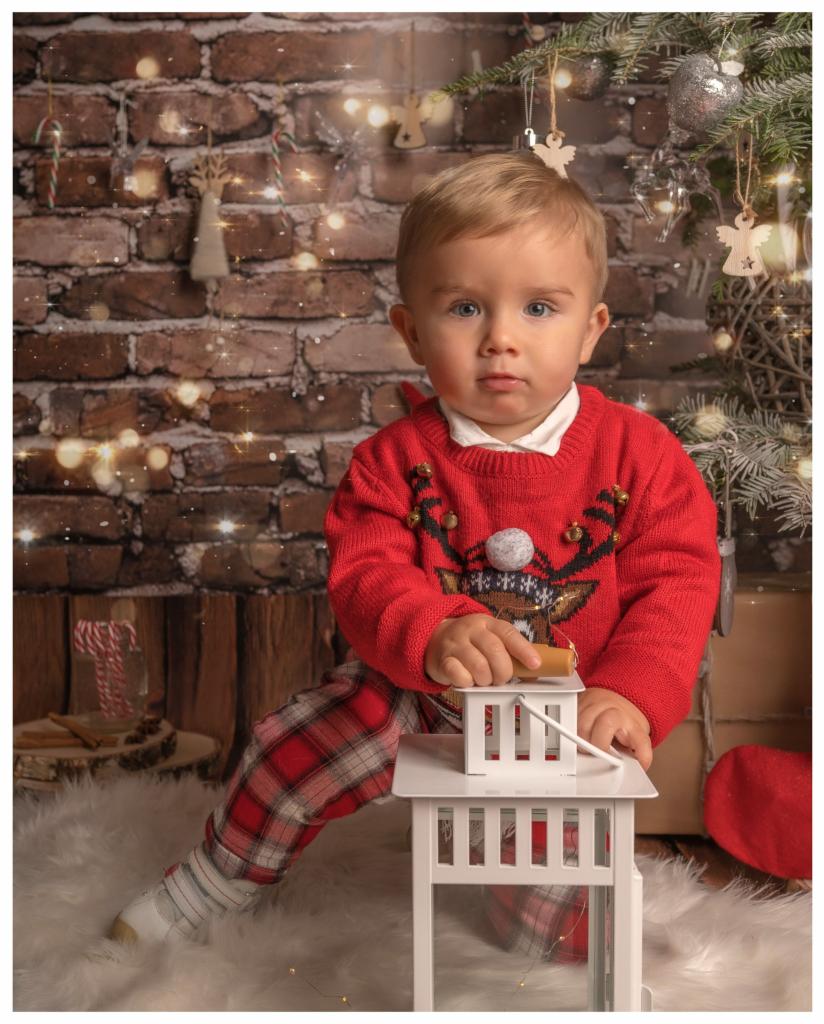 Mały chłopiec w świątecznym swetrze i kraciaste spodnie opiera się o białą latarenkę na tle ceglanej ściany z dekoracjami bożonarodzeniowymi i migoczącymi światełkami.